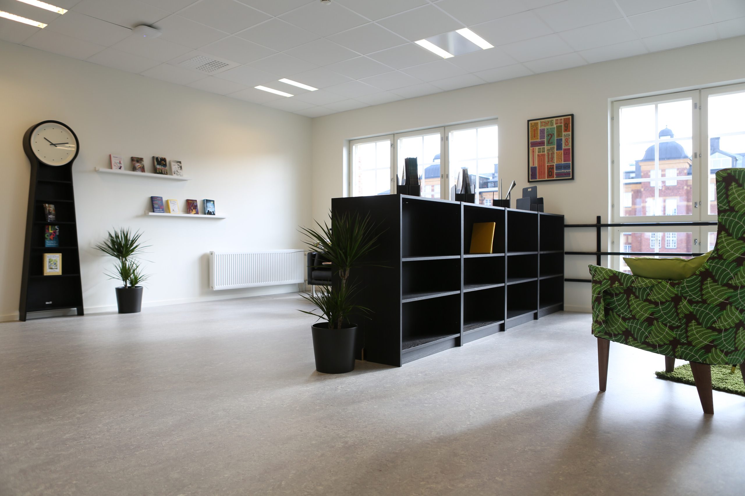 Ett ljust bibliotek eller läsrum med svarta bokhyllor, en klockformad bokhylla, gröna växter och en mönstrad fåtölj, med stora fönster som visar utsikt över omgivande byggnader.