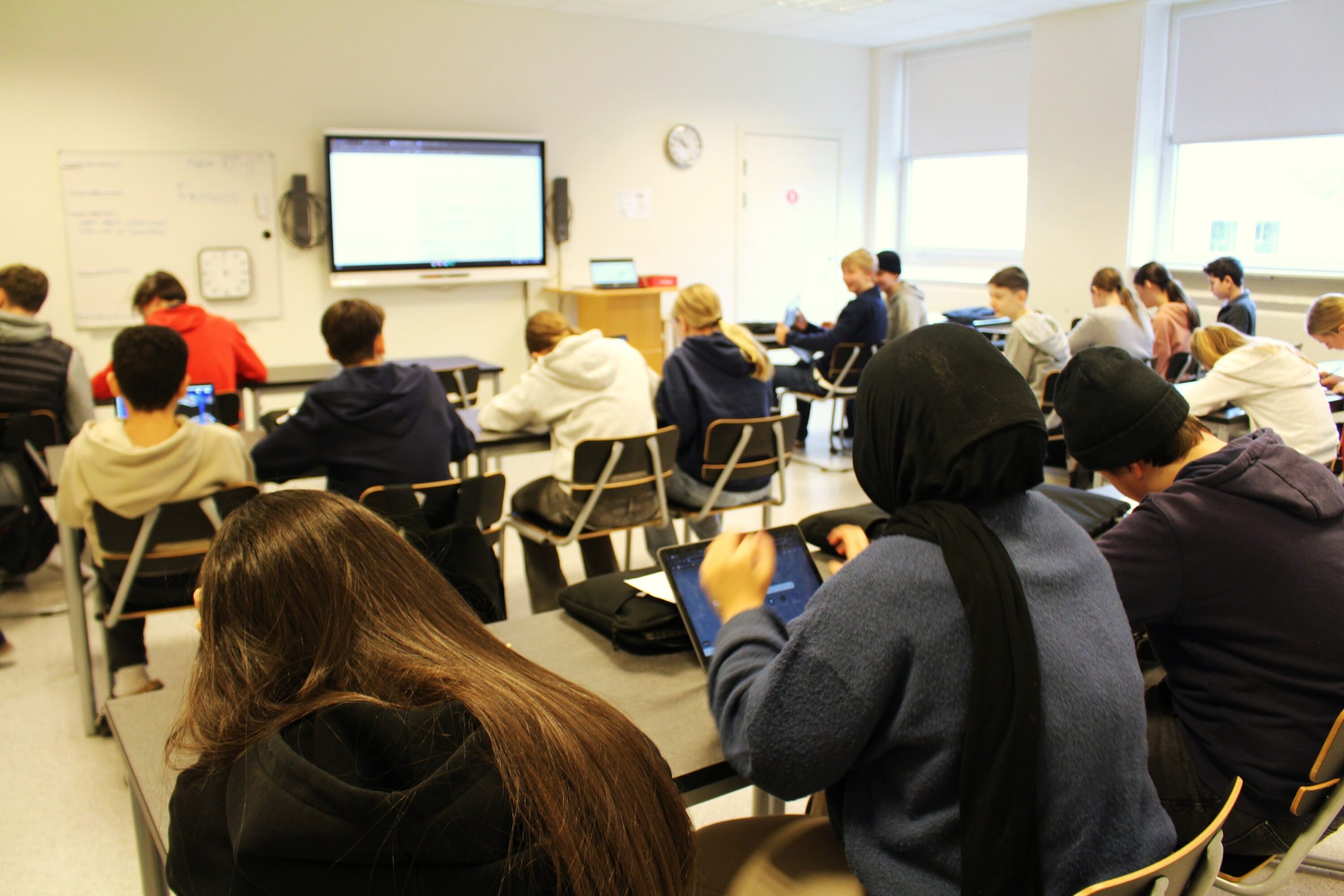 Elever sitter vid sina bänkar sedda bakifrån i ett klassrum, med en interaktiv skärm framme vid tavlan. Flera elever arbetar med surfplattor och laptops under lektionen.