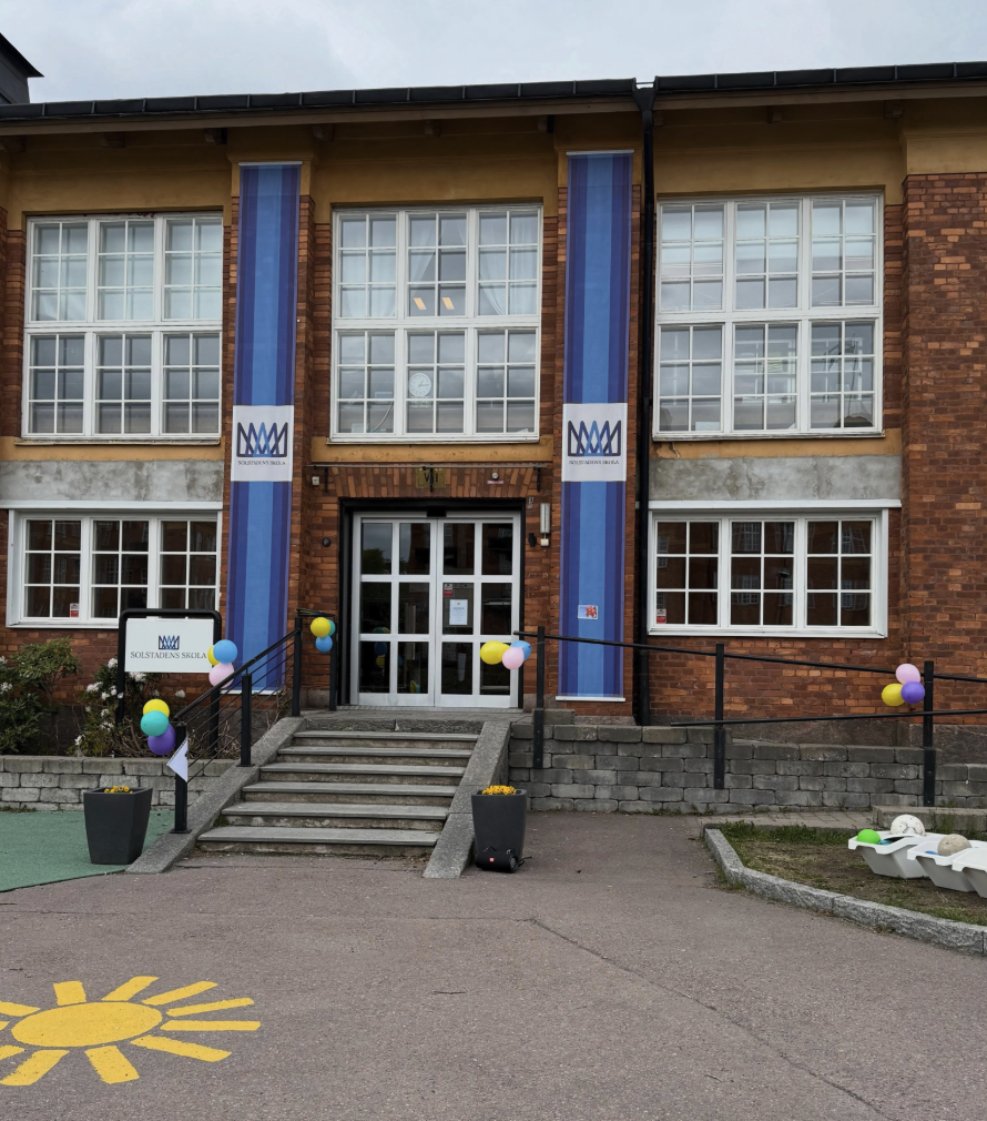Entrén till Solstadens skola, en tegelbyggnad smyckad med blåa banderoller och ballonger vid trappan, med en solmålning på asfalten framför entrén.