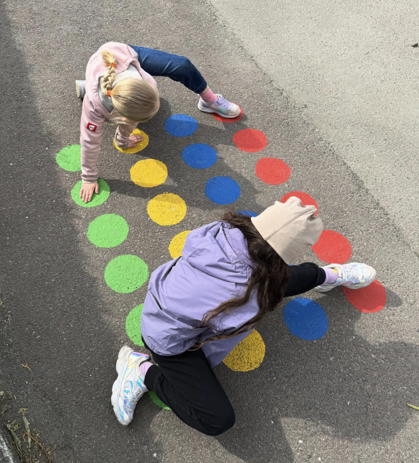 Två barn leker twister på en asfaltyta med färgglada målade cirklar i grönt, gult, blått och rött, fotograferade uppifrån.