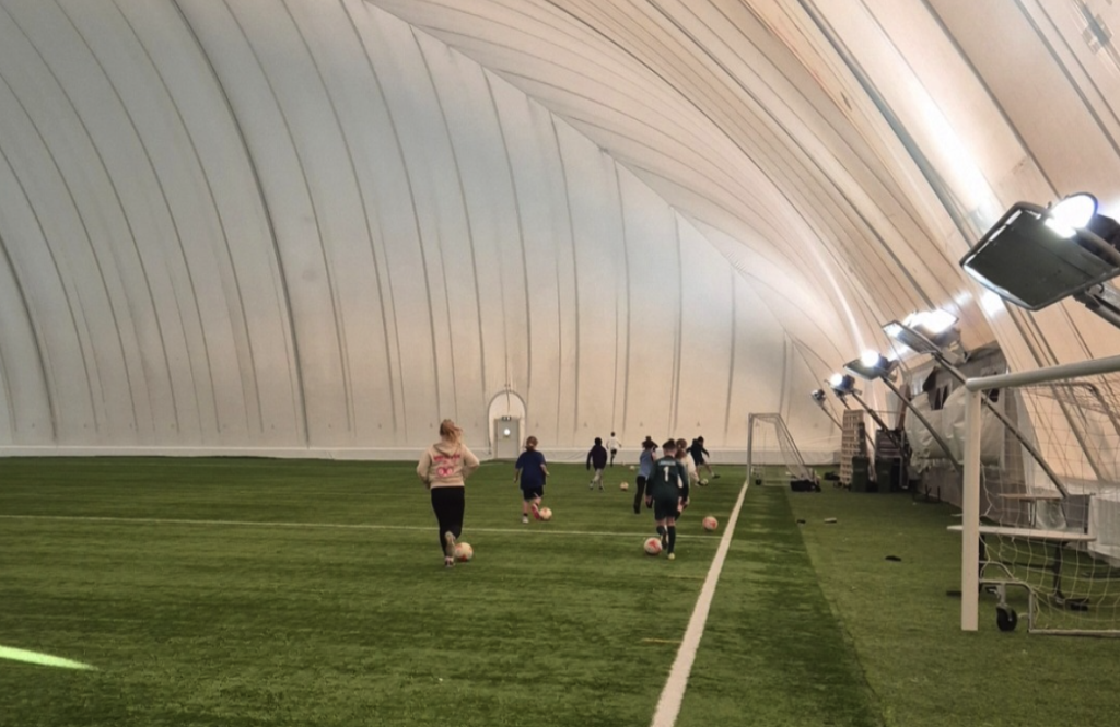Barn spelar fotboll med bollar på en konstgräsplan inne i en stor tälthall med valvformat tak.
