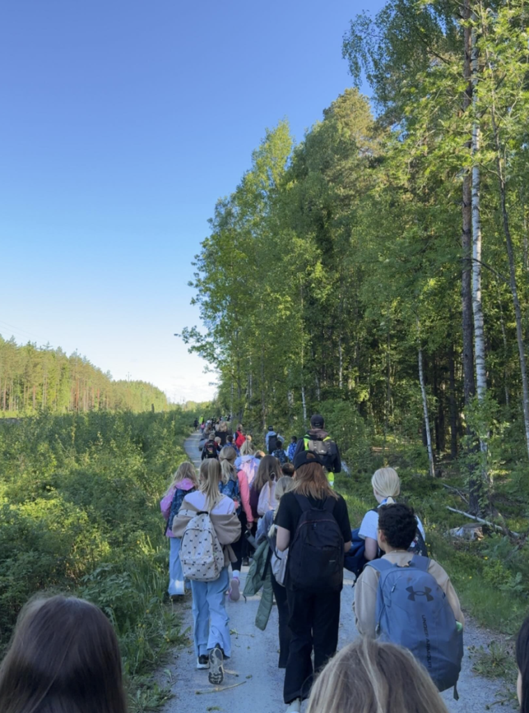 En lång rad ungdomar med ryggsäckar vandrar tillsammans på en grusväg längs skogsbrynet en klarblå dag.